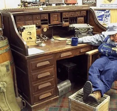 A figure in overalls at a roll-top desk with a telegraph key, typewriter and enamel mug.